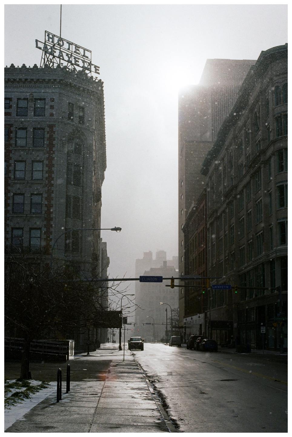 A street in a city is covered in snow with tall buildings on either side and a bright sky.