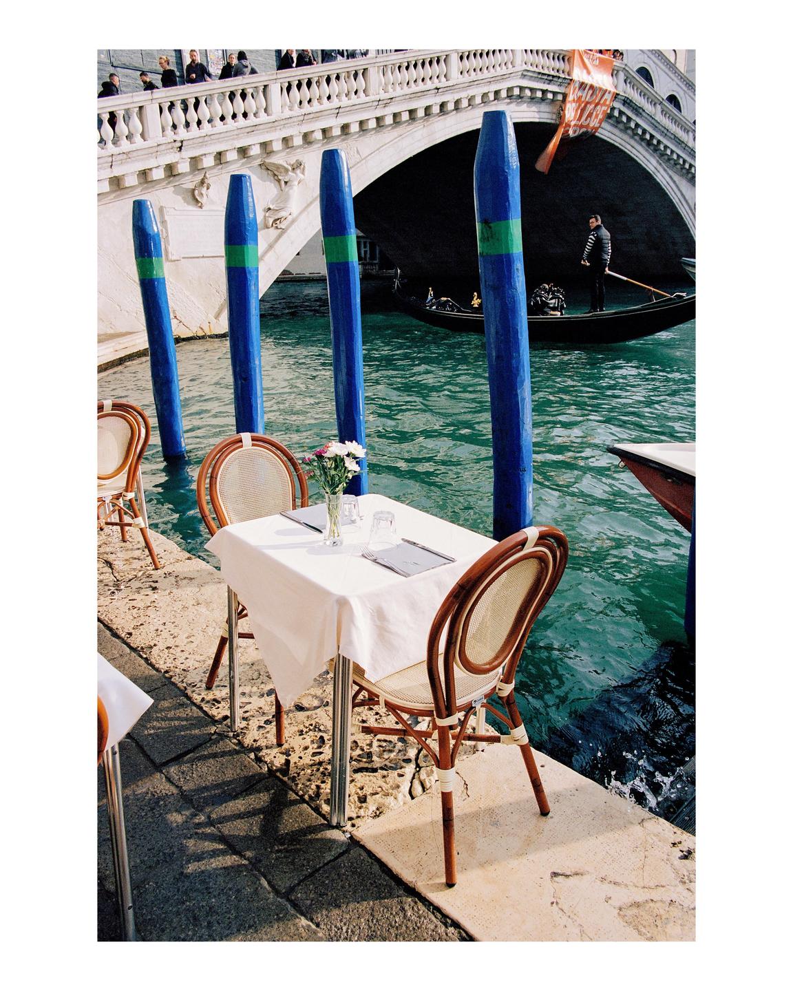 A table with two chairs sits beside a canal with a gondola passing under a bridge.