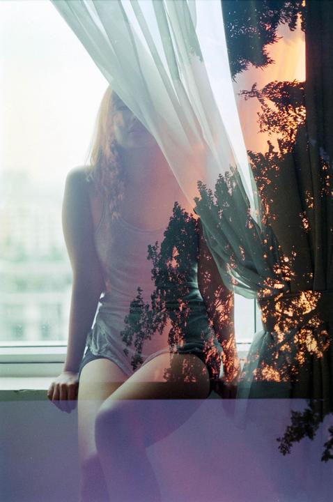 A woman sits by a window with a sheer curtain, overlaid with an image of trees against a sunset sky.