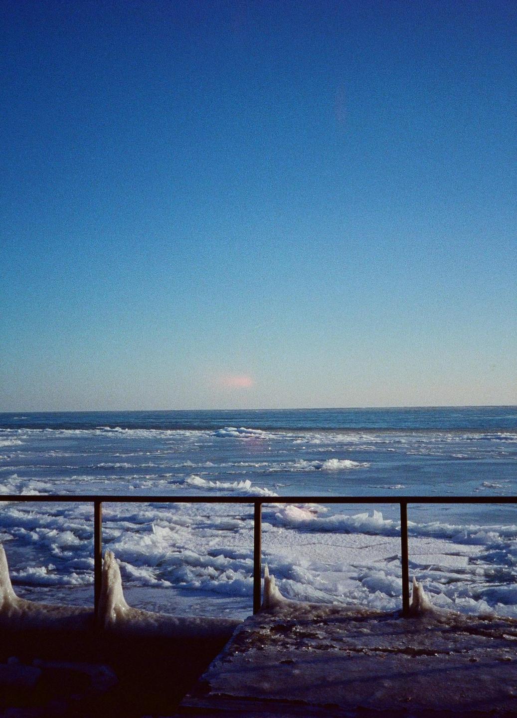 A metal railing covered in ice overlooks a frozen lake under a clear blue sky.