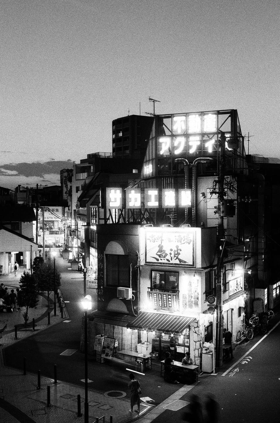 A street corner with buildings featuring neon signs and people walking is captured from an elevated perspective at night.