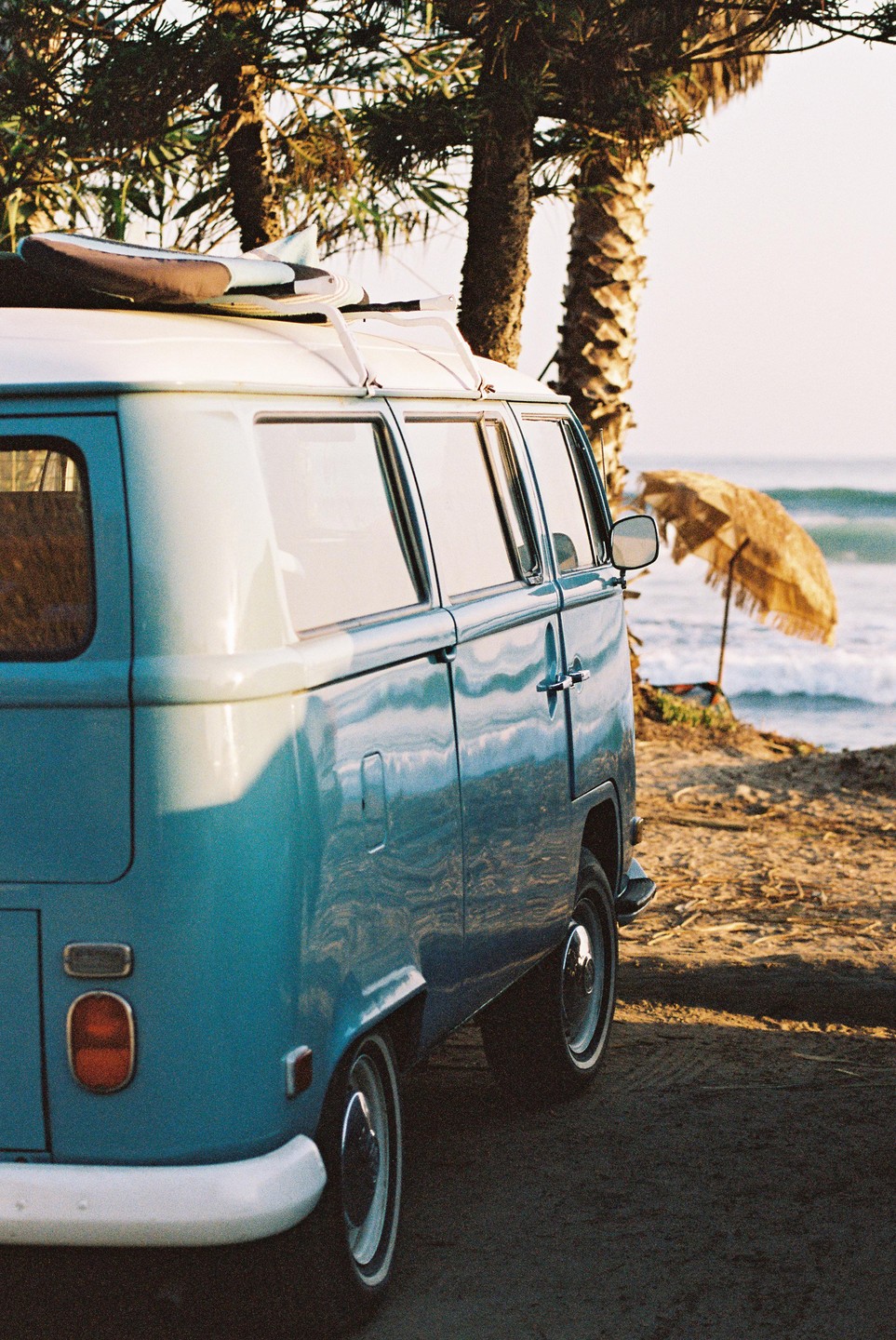 A blue van with a surfboard on its roof is parked on a dirt path next to a beach with palm trees and an umbrella.