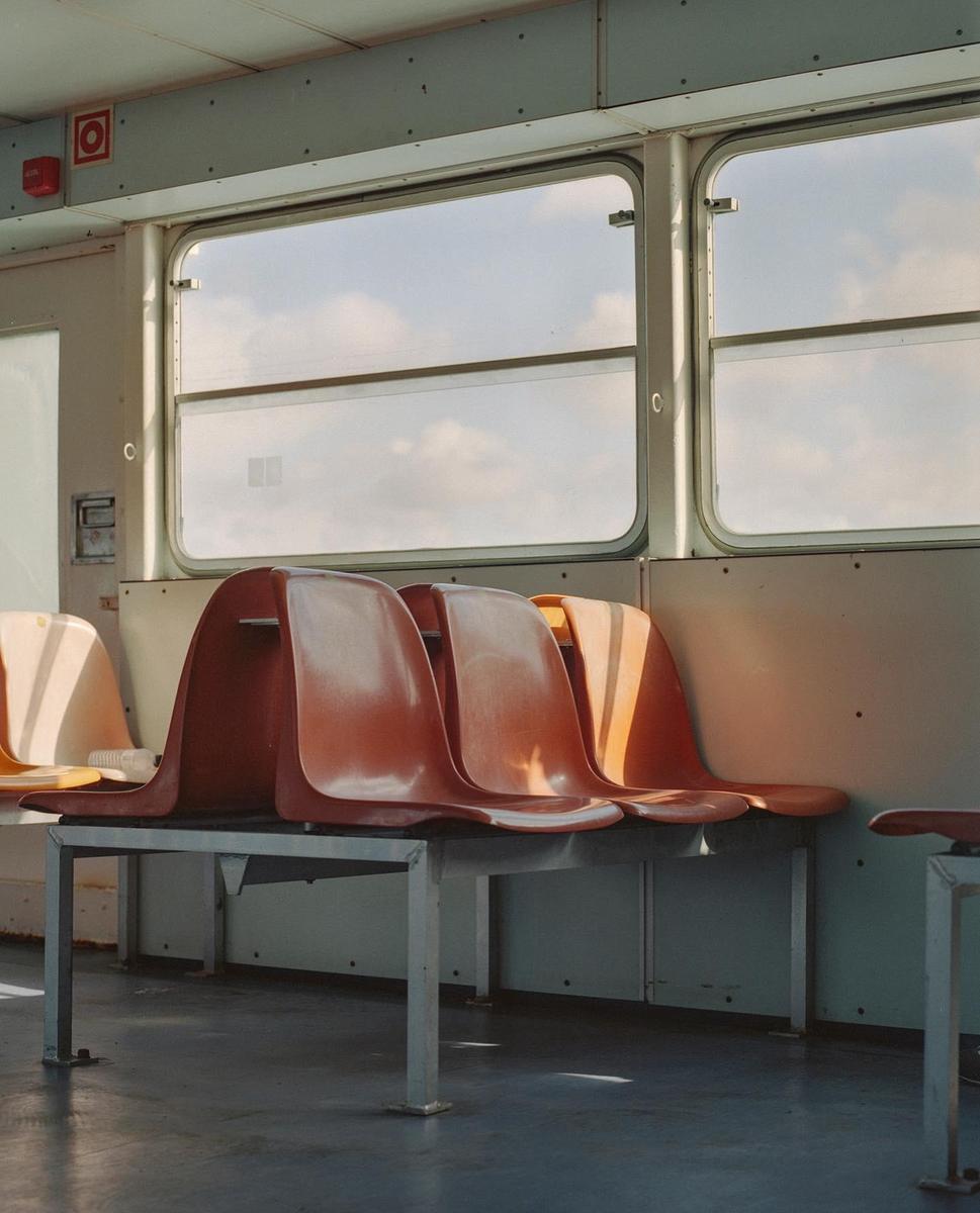 Empty orange seats are visible inside a ferry with windows showing a cloudy sky.