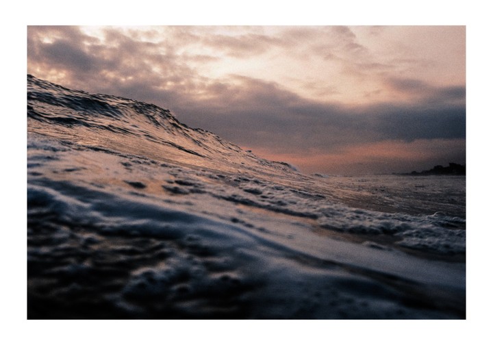 A wave crests under a cloudy sky at sunset.