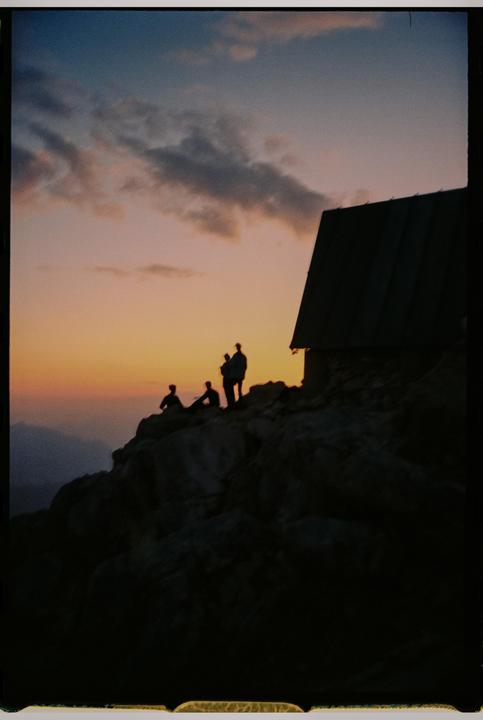 Silhouettes of people and a building are visible on a rocky mountain at sunset.