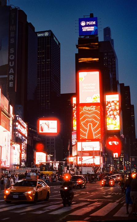 A street scene at night shows Times Square with bright billboards and vehicles on the road.