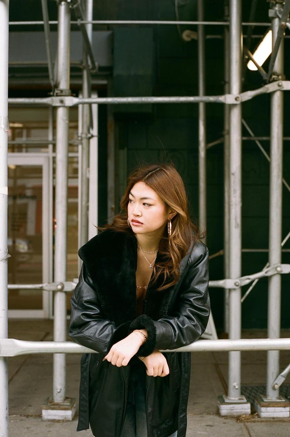 A woman in a black leather coat leans on a metal railing in front of a building.