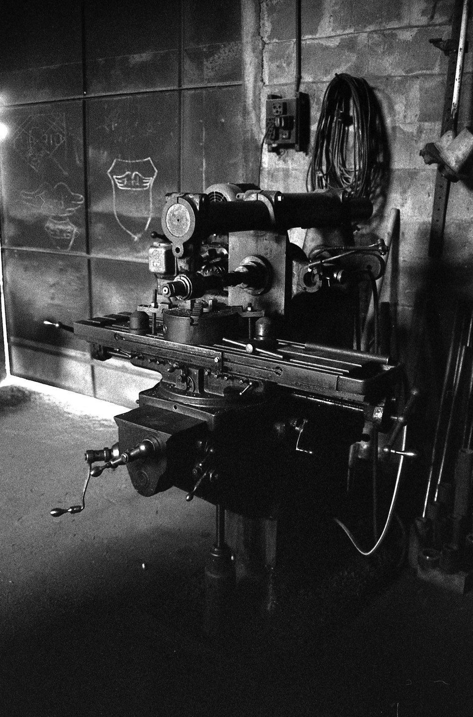 A large piece of machinery with various levers and parts is in a workshop with a wall covered in chalk drawings.