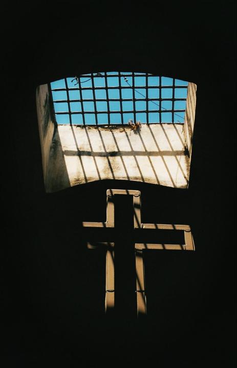 Sunlight streams through a barred window, casting a cross-shaped shadow onto a dark wall.