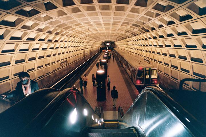 A subway station with a train and people on the platform is viewed from the top of an escalator.