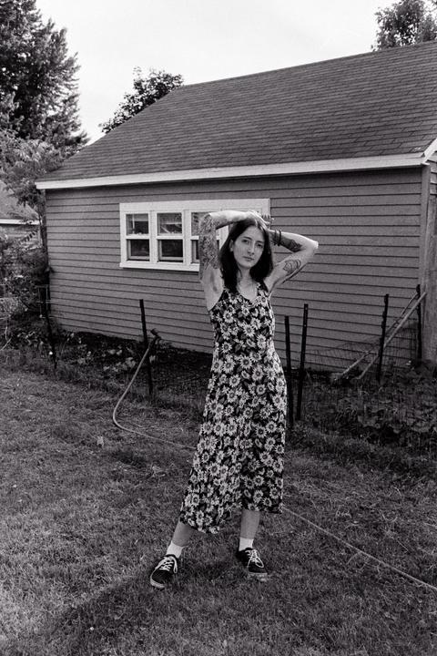 A woman with tattooed arms stands in a grassy yard in front of a house.