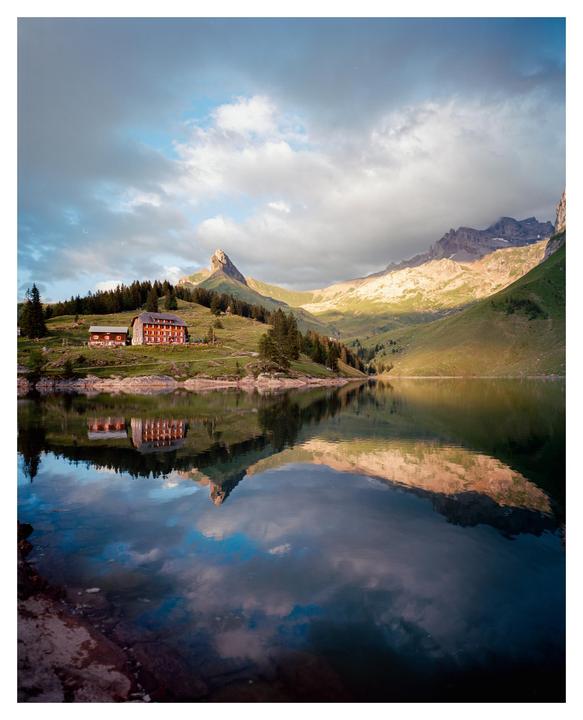 A red building sits on a grassy hill next to a lake, with mountains and clouds reflected in the water.