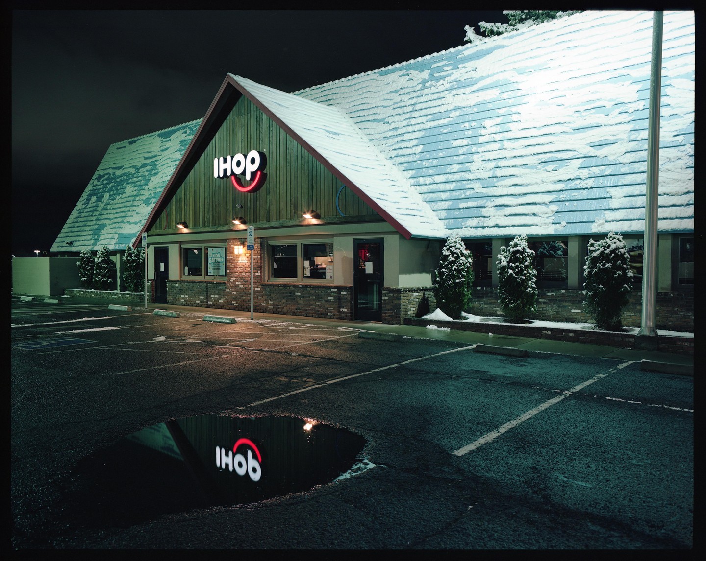An IHOP restaurant with a snow-covered roof is seen at night, with its neon sign reflected in a puddle in the parking lot.