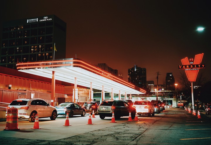Cars are lined up at night under a brightly lit drive-through canopy with a large sign for The Varsity.