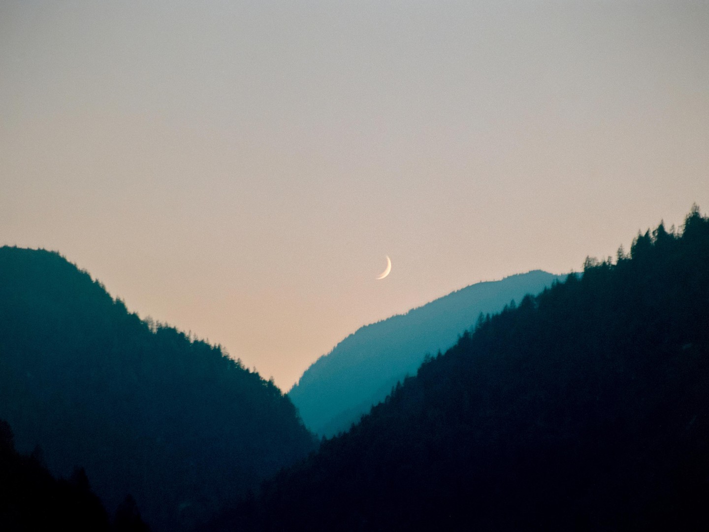 A crescent moon hangs in the sky above two dark, forested mountain ridges.