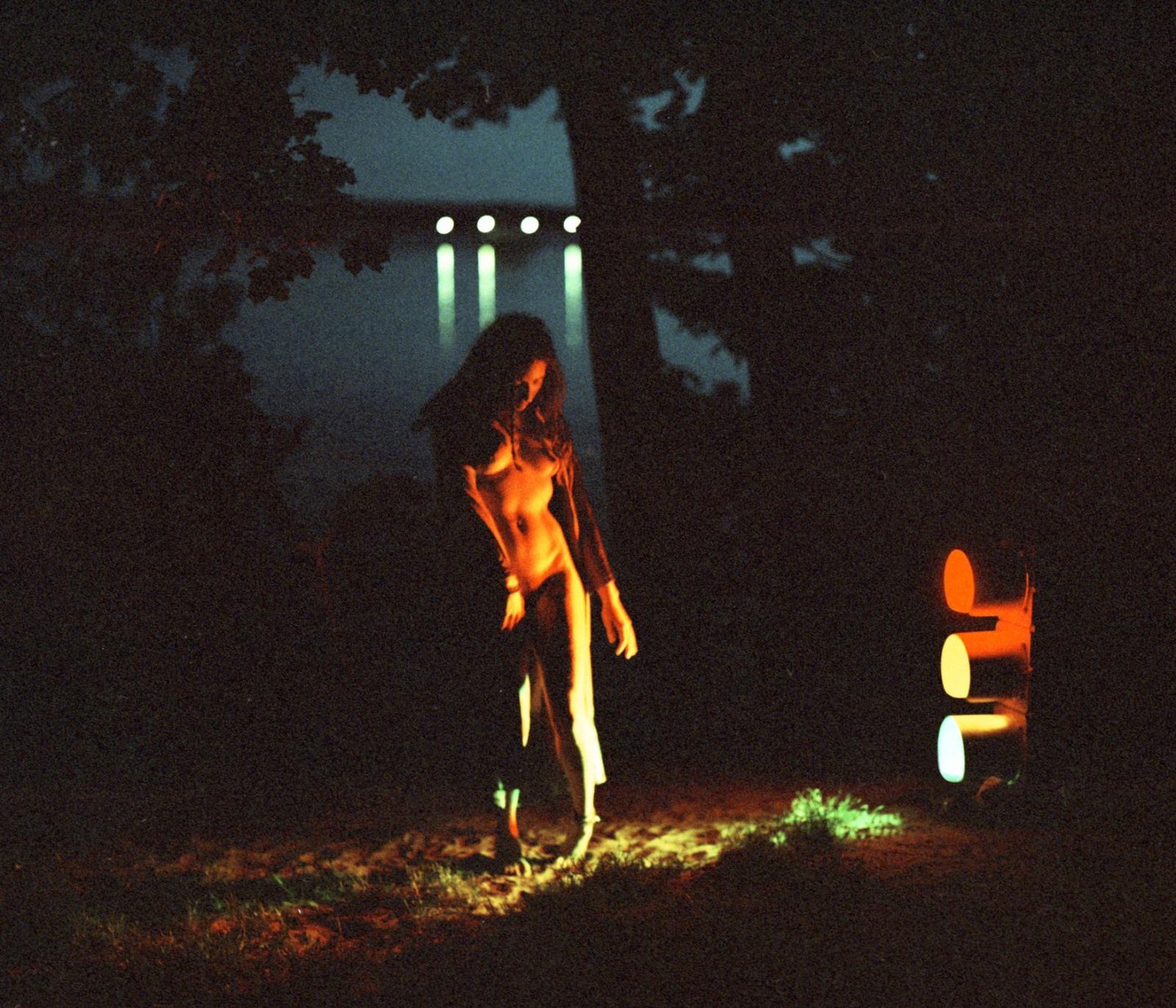 A woman stands in the dark illuminated by a traffic light and other lights in the distance.