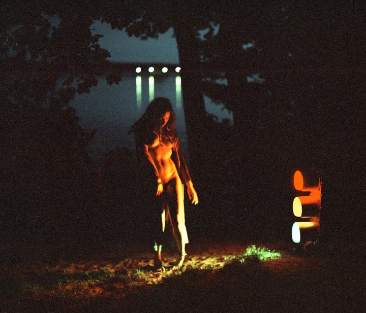 A woman stands in the dark illuminated by a traffic light and other lights in the distance.