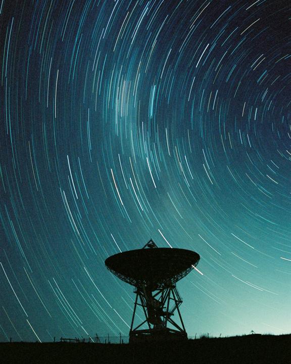 A radio telescope is silhouetted against a night sky filled with star trails.