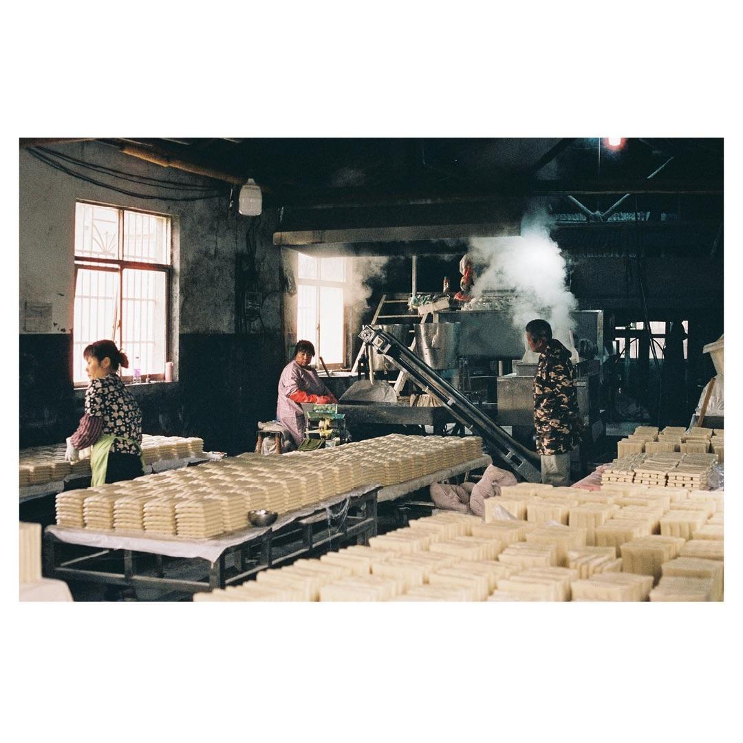 Three people work in a rice cake factory with large windows and machinery producing steam.
