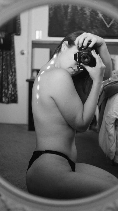 A woman kneels on the floor, taking a self-portrait in a mirror, with light spots on her back.