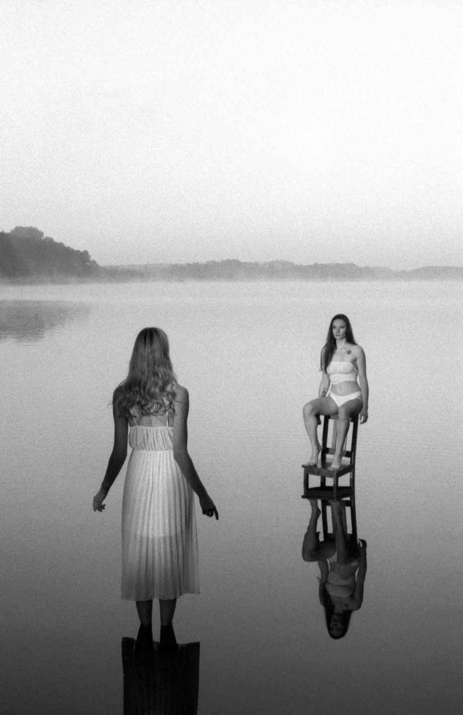 A woman in a dress stands on a submerged platform facing another woman in a bikini sitting on a chair in a misty lake.