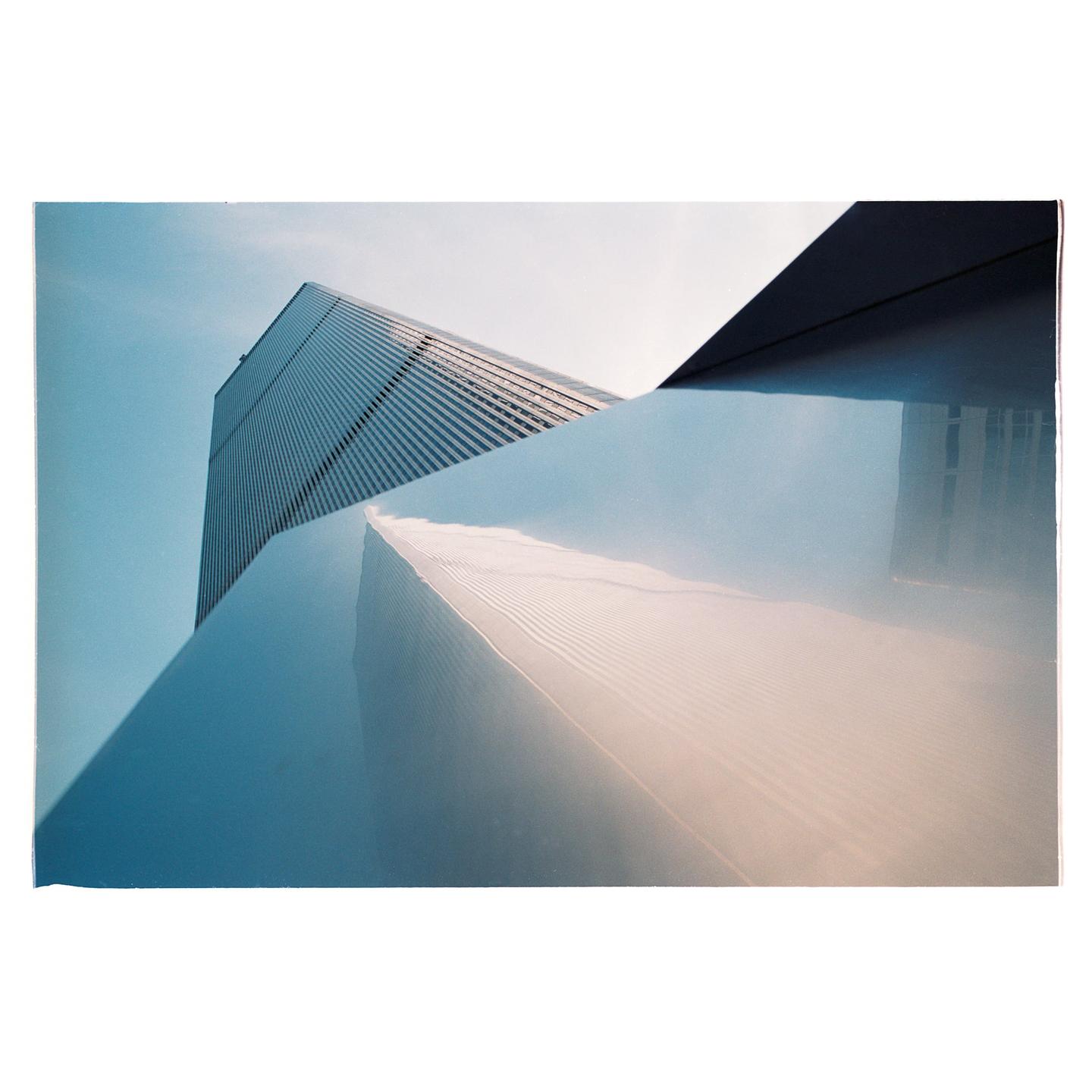 A tall building with many windows is reflected in a shiny, angled surface under a blue sky.