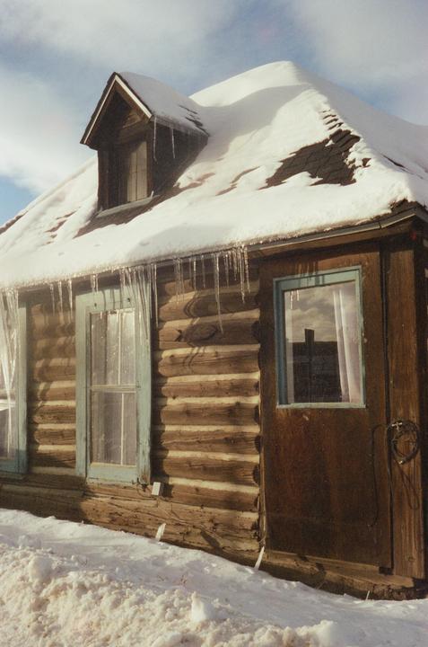 A log cabin with a snow-covered roof and icicles hanging from the eaves is shown in winter.