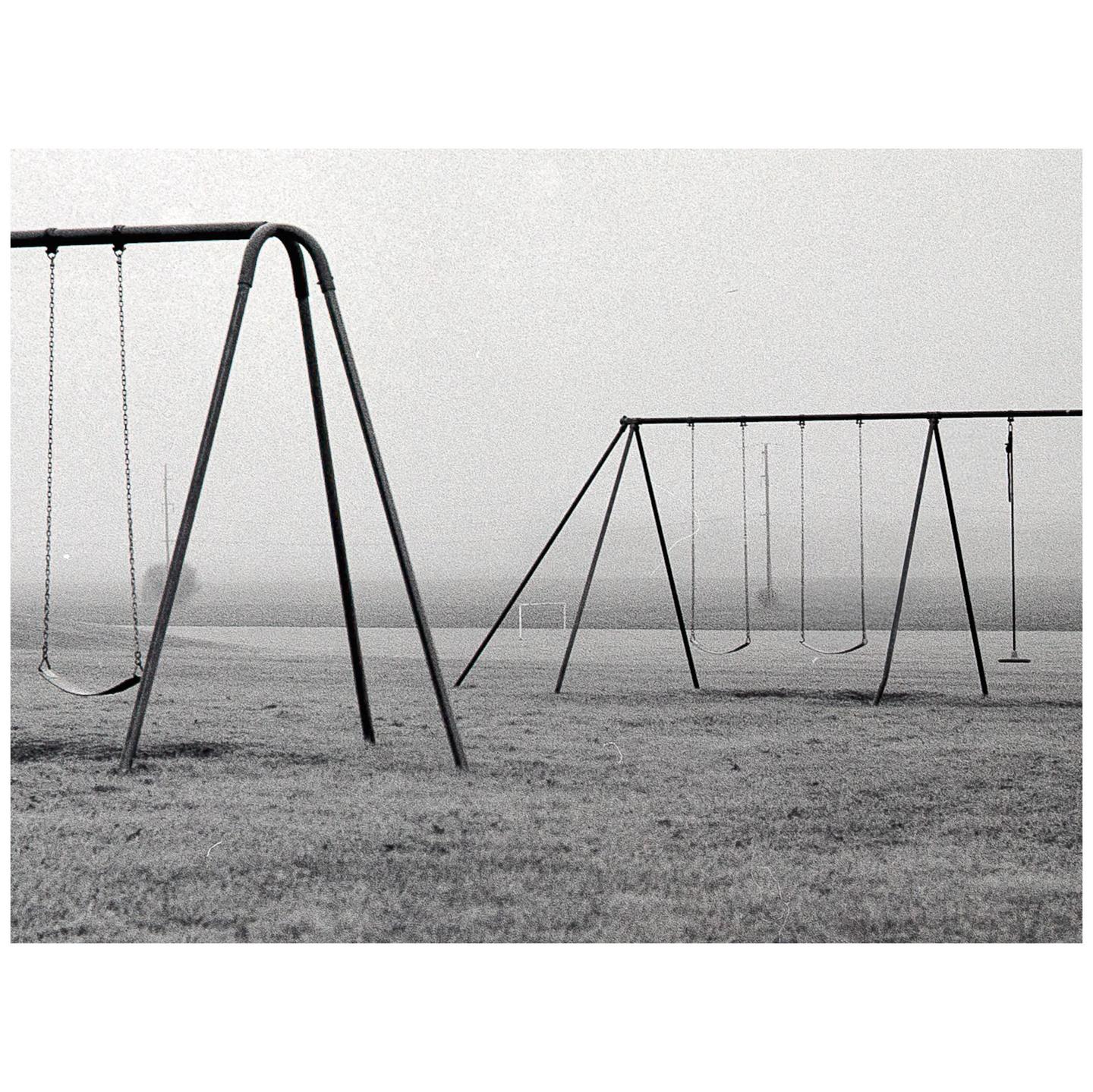 Two swing sets stand on a grassy field under a hazy sky.
