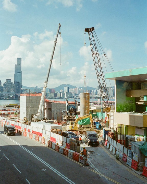 Two cranes are visible at a construction site next to a road with cars and a city skyline in the background.