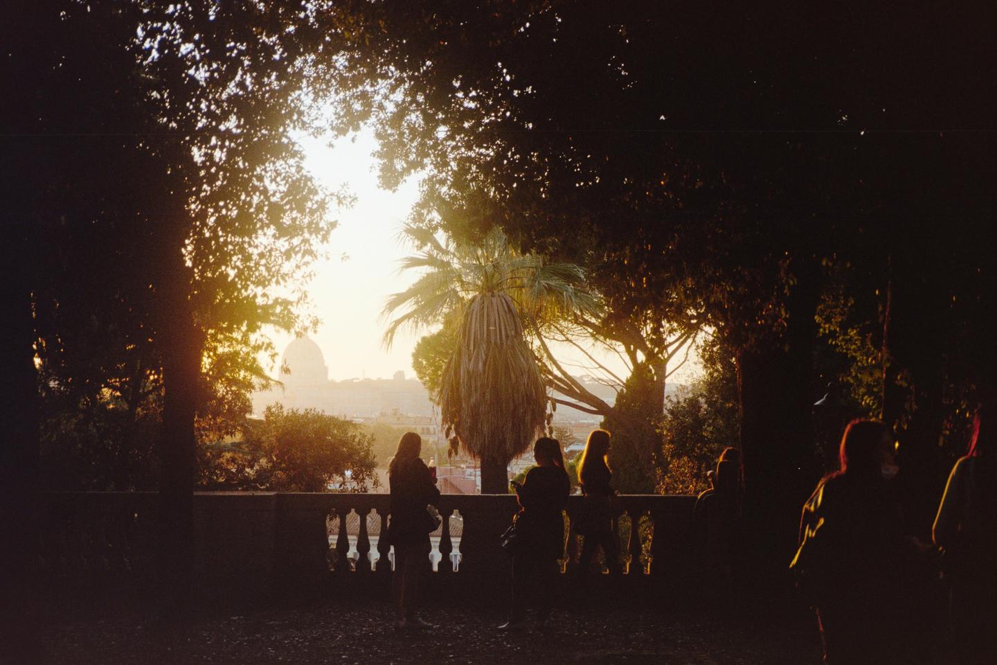 People stand on a balcony overlooking a city with a large dome and palm trees at sunset.