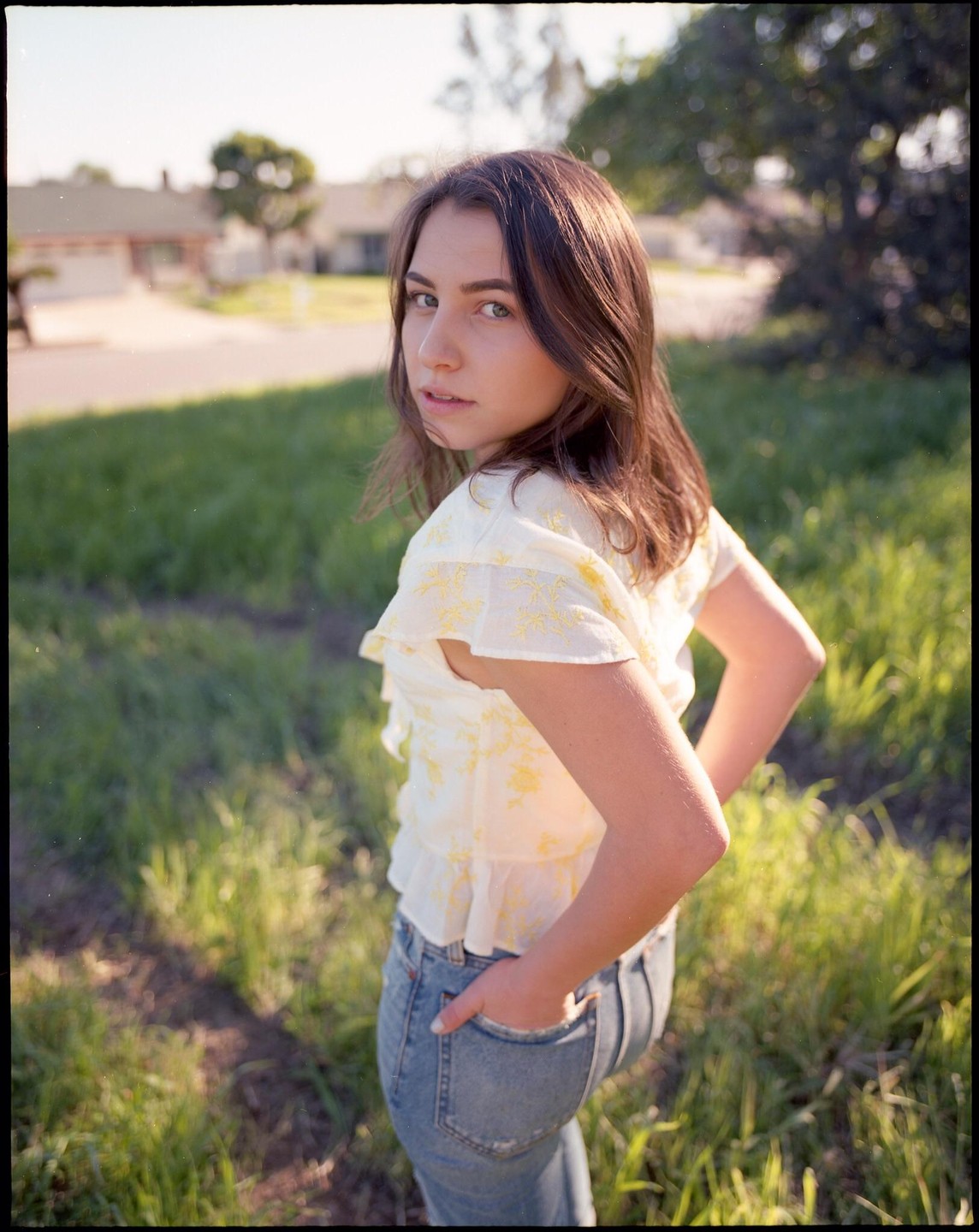 A young woman with long hair looks over her shoulder at the viewer while standing in a grassy field.