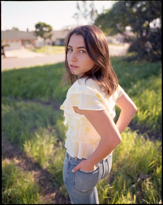 A young woman with long hair looks over her shoulder at the viewer while standing in a grassy field.