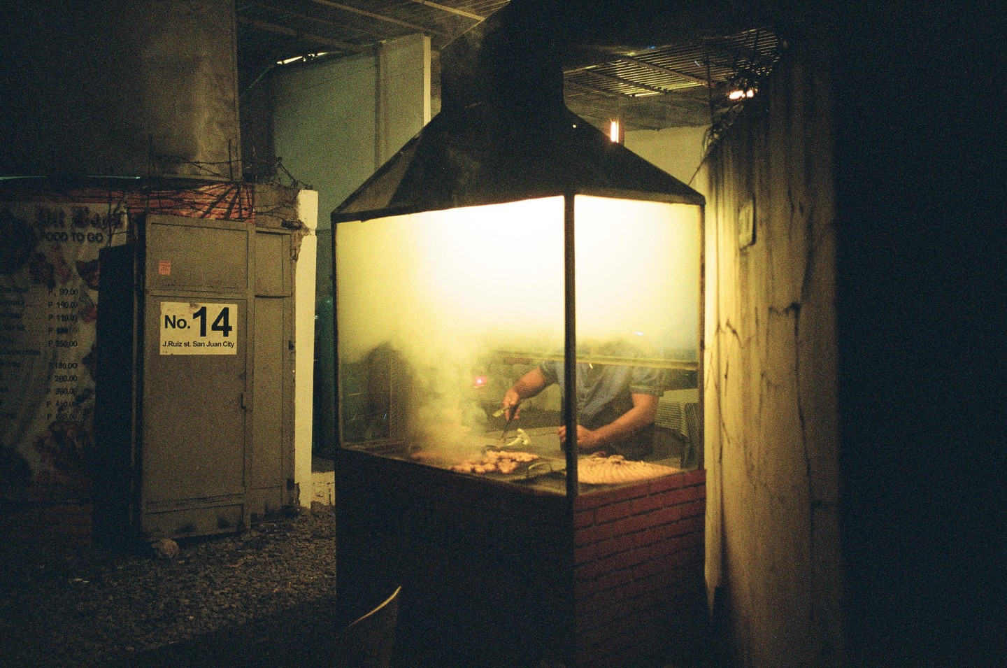 A man cooks food on a smoky grill inside a brightly lit glass enclosure at night on a street in San Juan City.