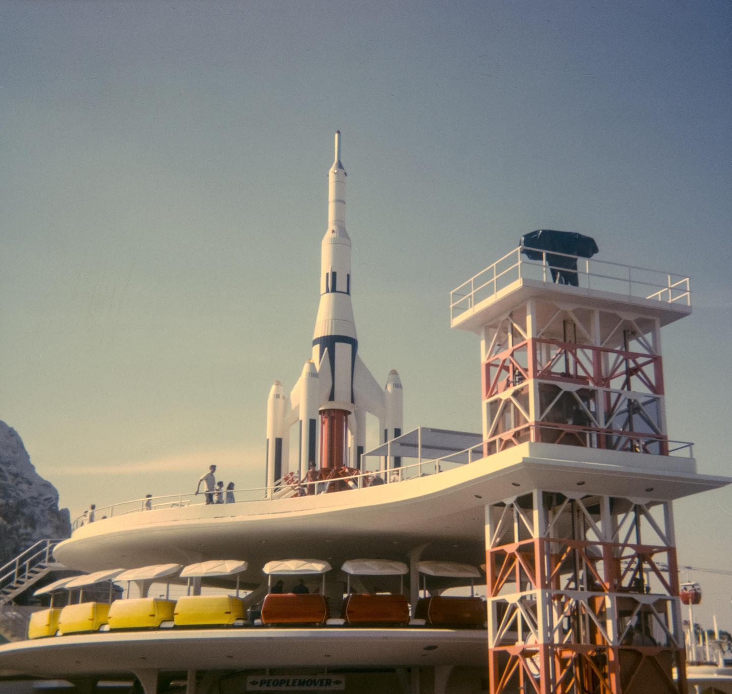 A large white rocket stands next to a building with a people mover ride and a tall tower.