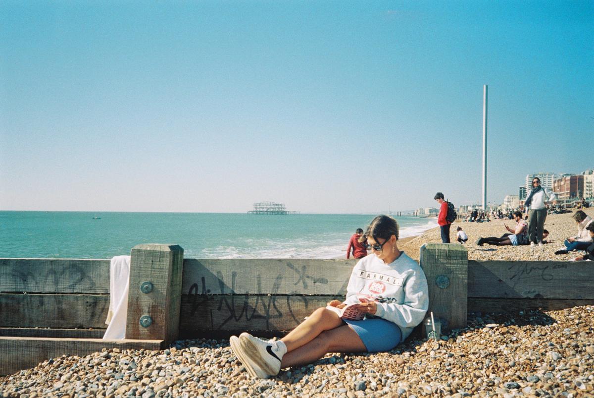 A woman sits on a pebble beach reading a book, with the ocean and a pier in the background.