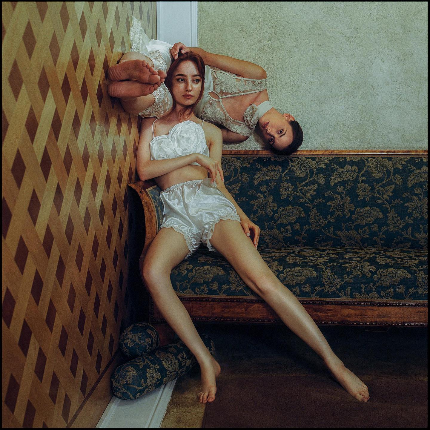 Two women in lingerie pose in a room with patterned wallpaper and a floral sofa.