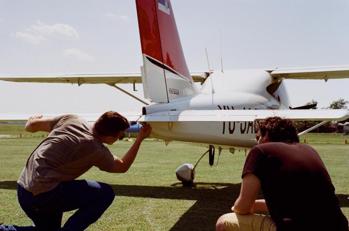 Two men are performing a pre-flight check on a small white airplane with a red tail.