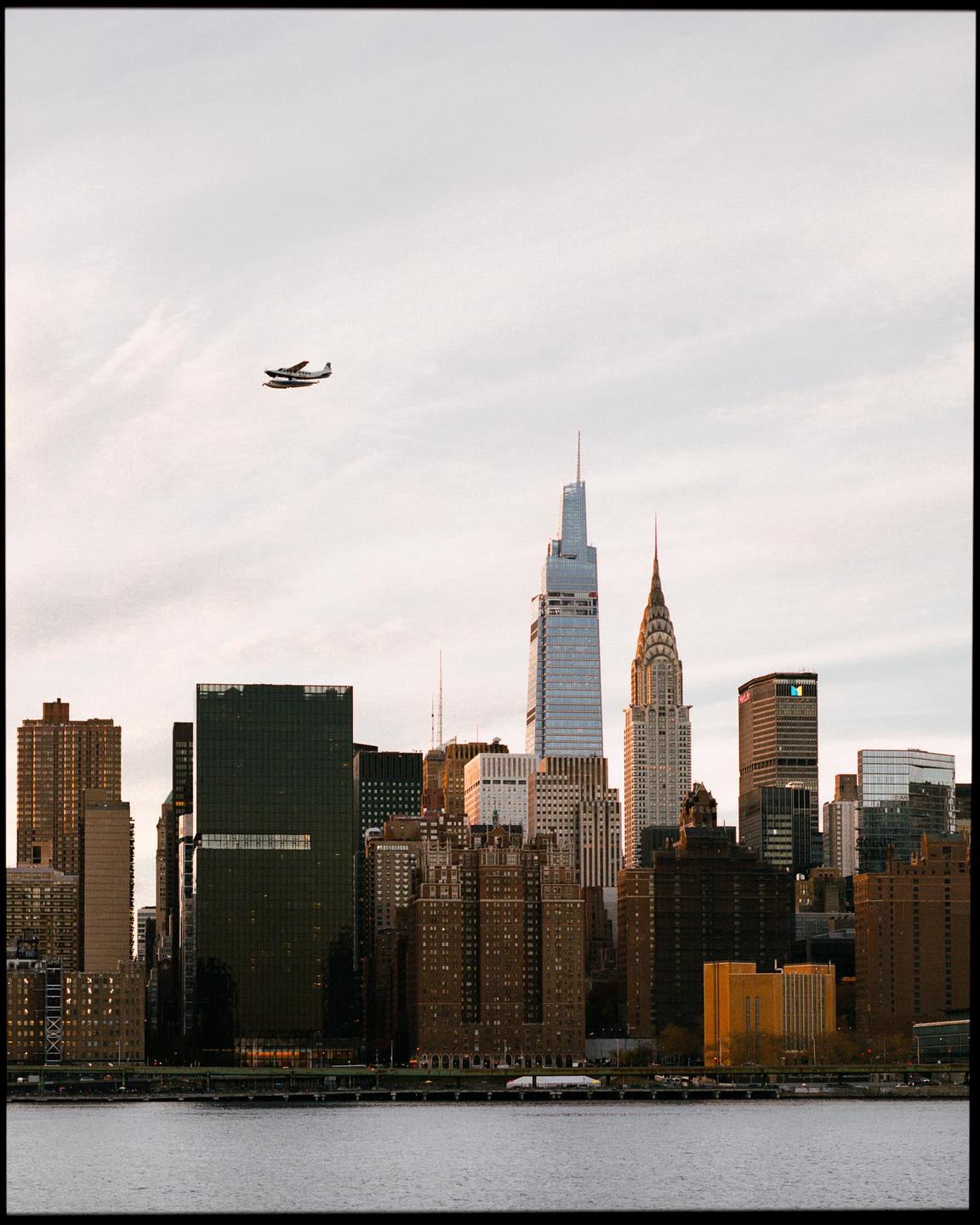 A seaplane flies over a city skyline with a body of water in the foreground.