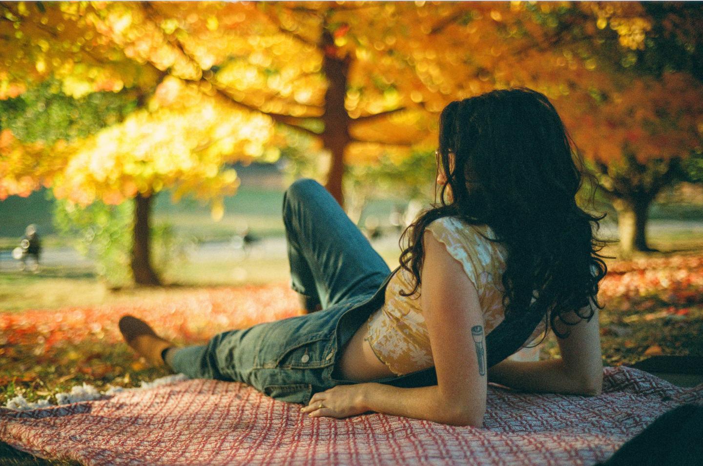 A woman is lying on a blanket in a park with autumn foliage.