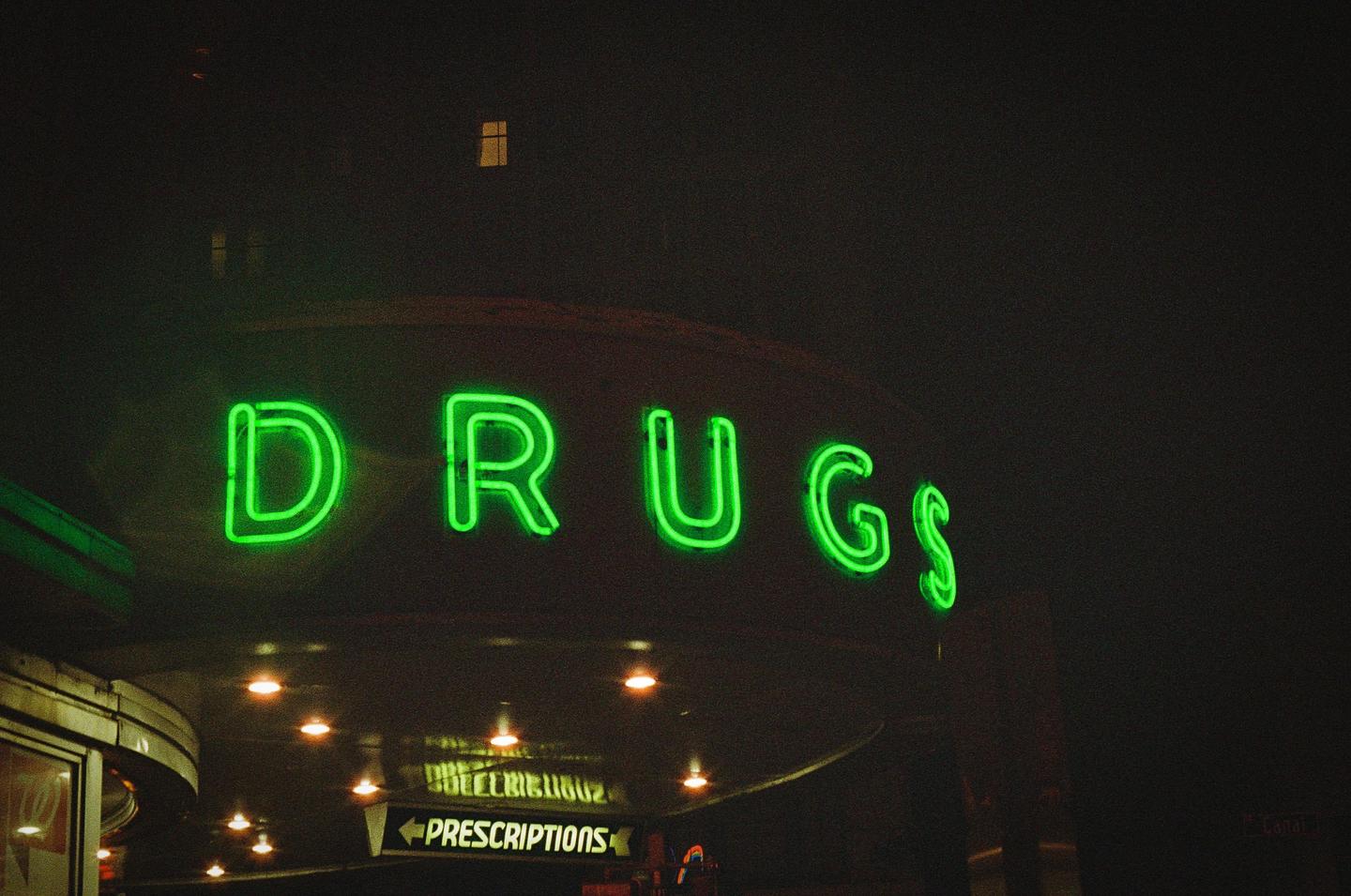 A neon green "DRUGS" sign is illuminated at night above an entrance with a "PRESCRIPTIONS" sign.