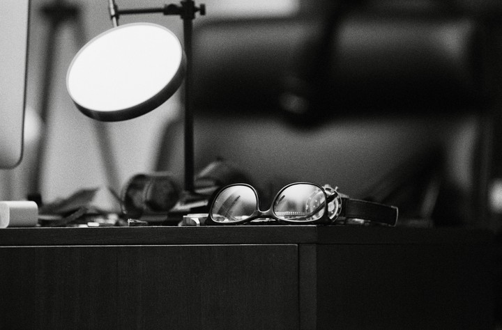 A pair of eyeglasses rests on a dark surface next to a lamp and other items.