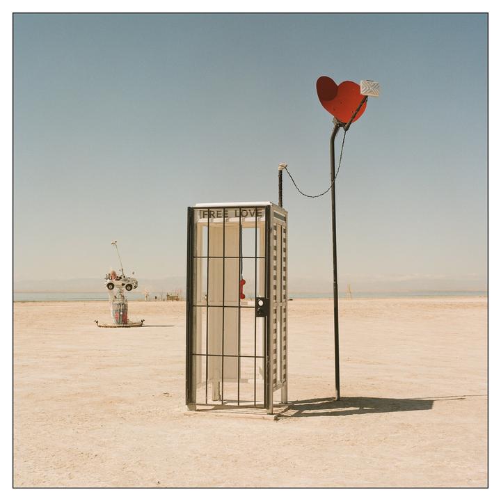A phone booth with a red telephone stands in a desert landscape next to a pole with a red heart and a white envelope.