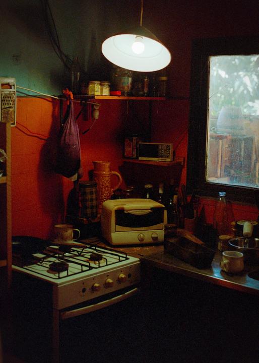 A kitchen with a stove, toaster oven, and various items on shelves and a counter is illuminated by a hanging lamp and light from a window.