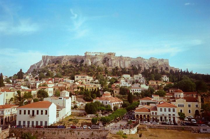 A wide view shows the Acropolis of Athens towering over the Plaka neighborhood, with many buildings and trees in the foreground.