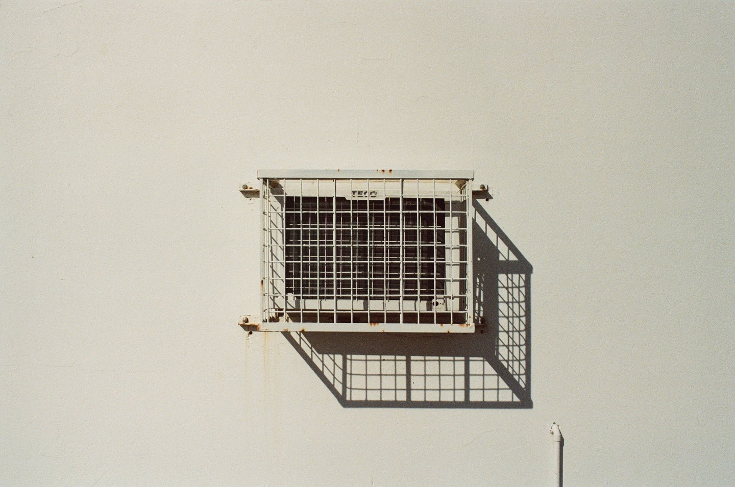 A white air conditioning unit with a grid pattern is mounted on a plain white wall, casting a strong shadow.