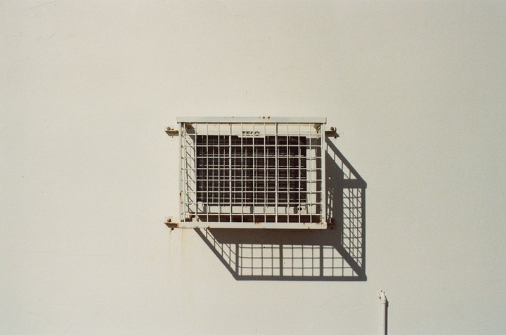 A white air conditioning unit with a grid pattern is mounted on a plain white wall, casting a strong shadow.