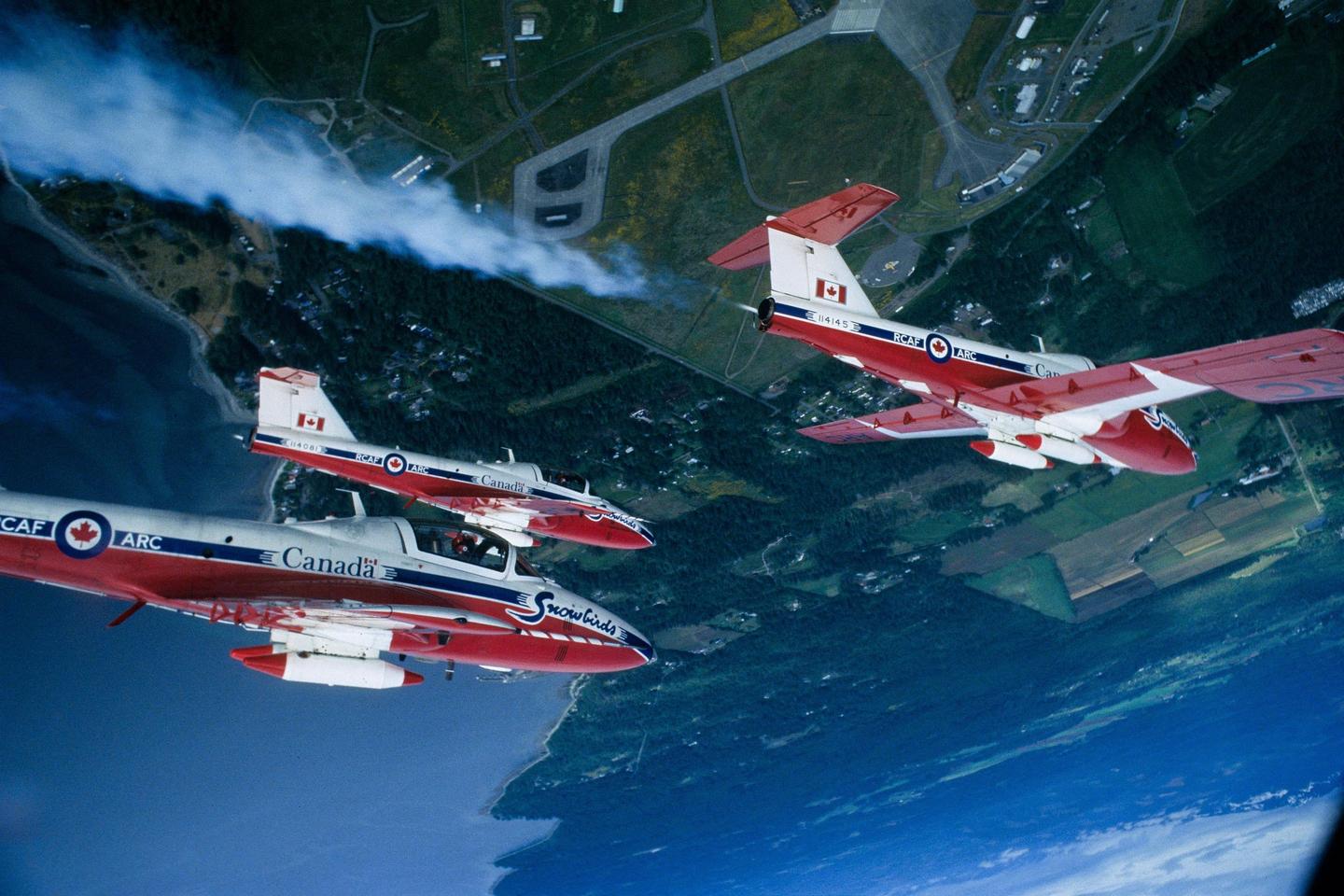 Three red and white Snowbirds jets fly in formation over a coastal landscape with an airport visible.