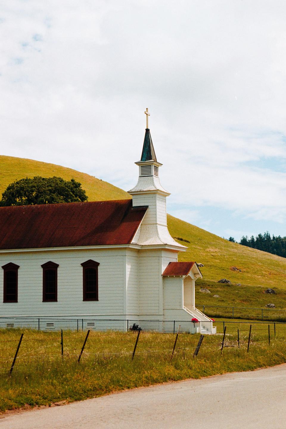 A white church with a red roof and steeple stands beside a road, with green hills and trees in the background under a cloudy sky.
