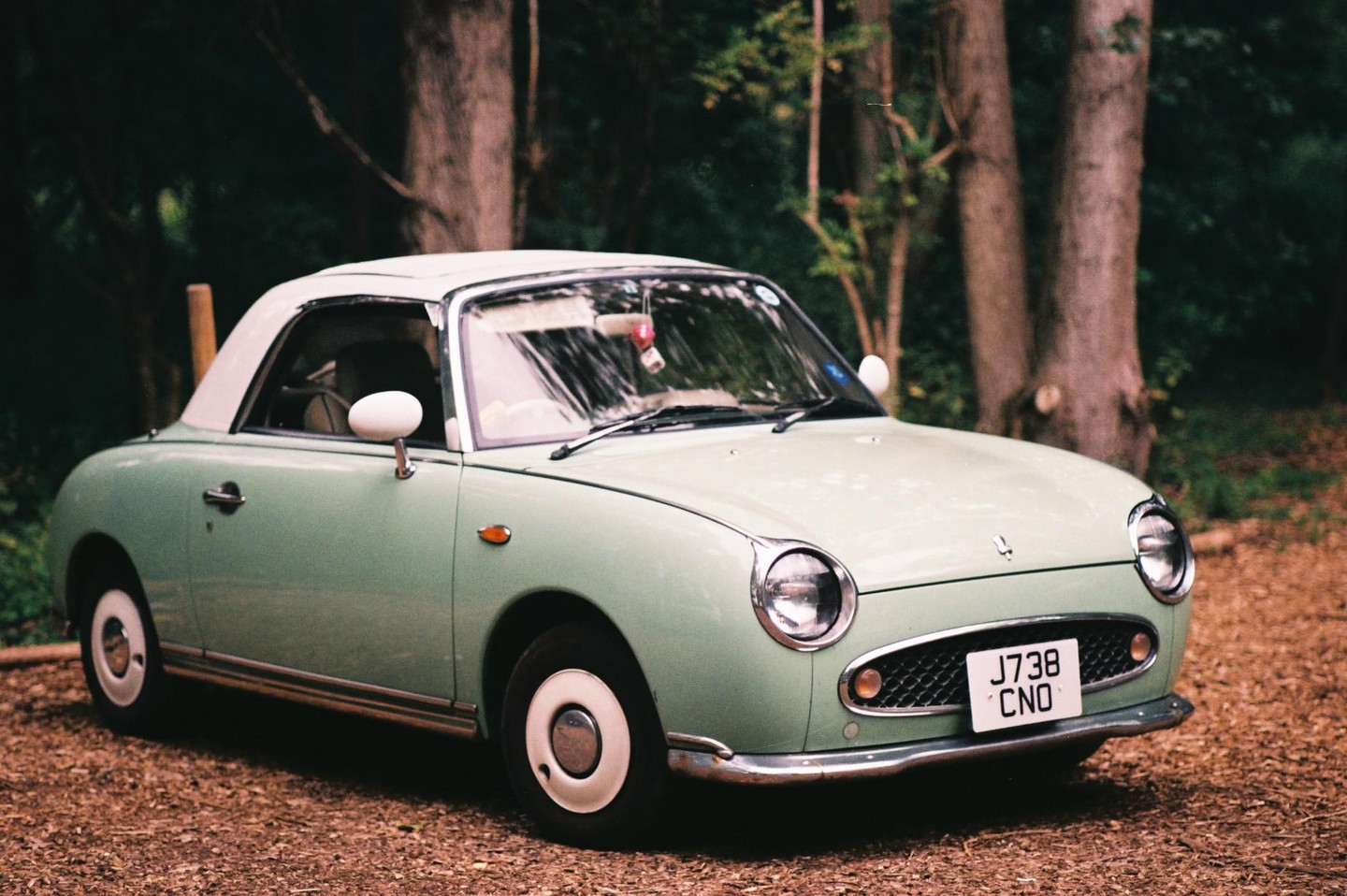 A light green vintage car with a white roof is parked on a dirt path surrounded by trees.