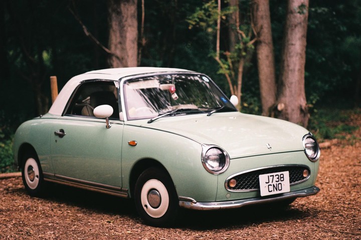 A light green vintage car with a white roof is parked on a dirt path surrounded by trees.